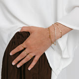 Model wearing an 18k gold-plated jewelry set featuring a ring stack with the Mughal Bloom Bezel Ring on the index finger, Intertwined Bezel Ring on the middle finger, and North Star Bezel Ring on the ring finger, paired with a wrist stack featuring the Tennis Name Bracelet with custom Arabic calligraphy alongside a pavé crystal tennis bracelet and a delicate charm bracelet.