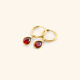 Alt text: The Garnet Stone Huggie Hoops by Nominal, crafted with an 18k gold-plated finish featuring smooth hoop bands accented with dangling teardrop red garnet gemstone charms, displayed on an off-white background.