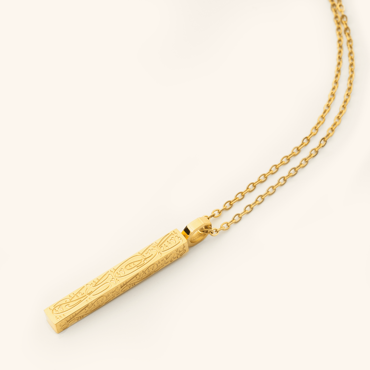Photo of an 18k gold plated necklace that features the Arabic Calligraphy of 'with every hardship comes ease' that is engraved intricately all along this timeless bar pendant.