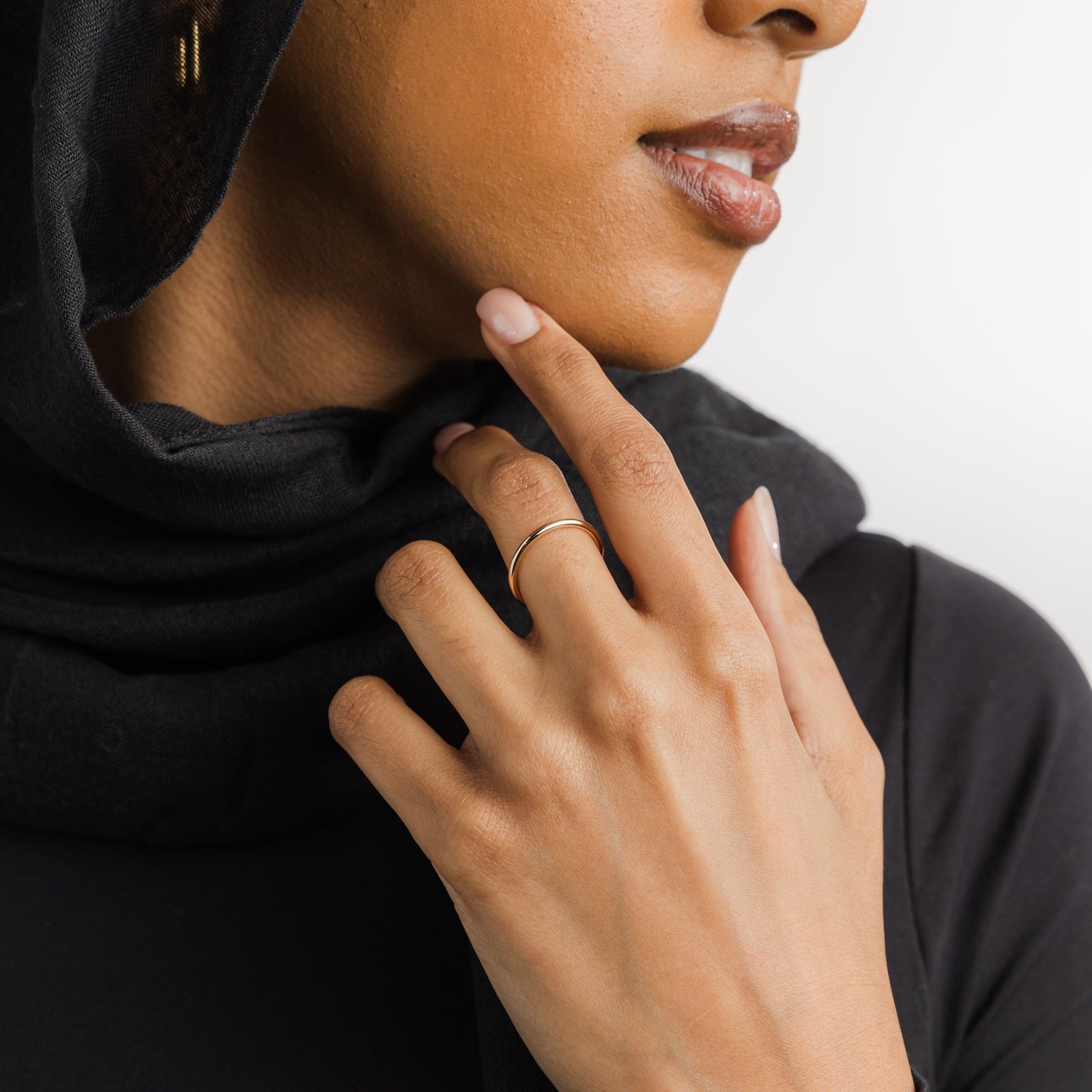 Woman wearing a thin gold band on her finger, hand touching her chin, styled with a black hijab.