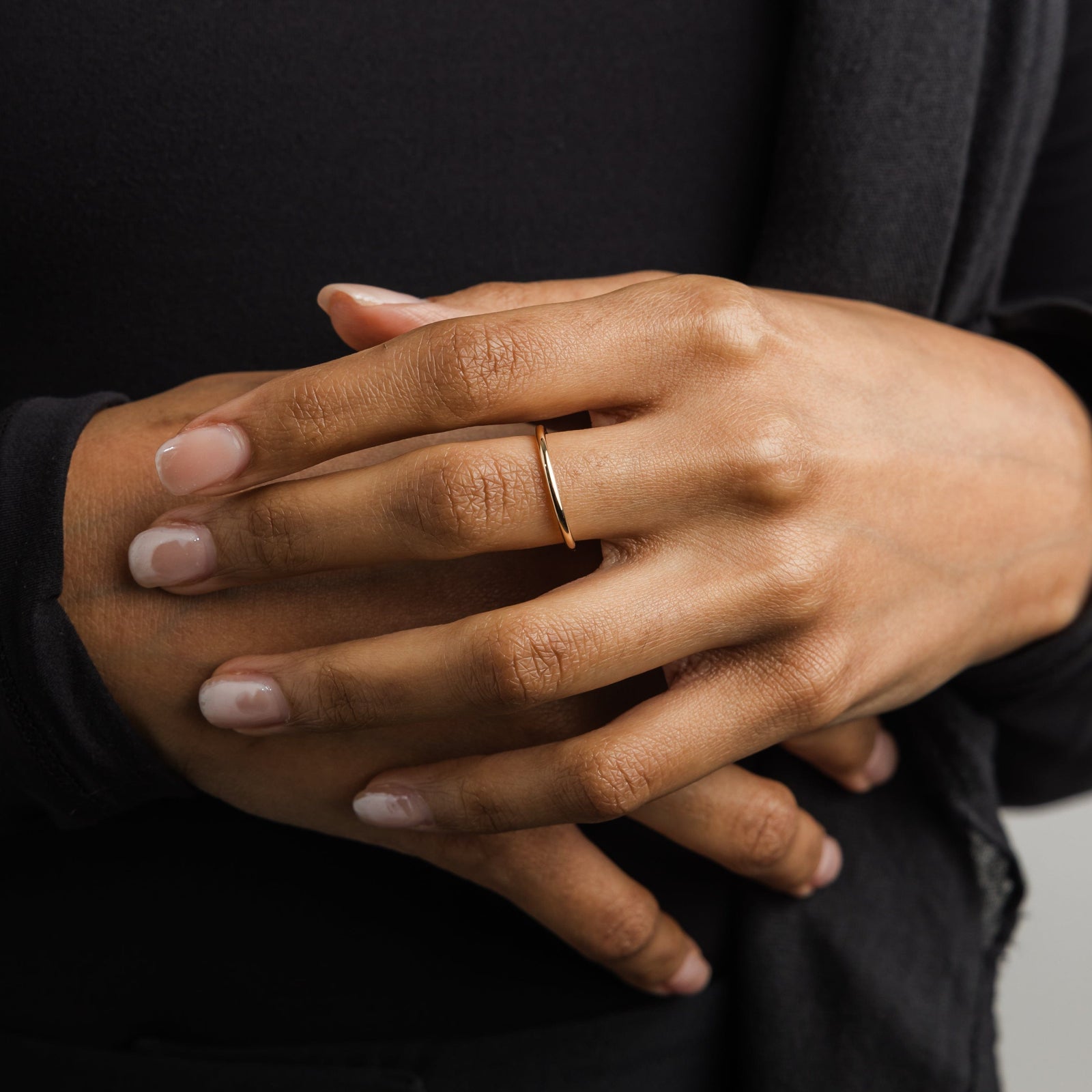 Woman’s hands, wearing a thin gold band on her finger, styled with neutral nails and black outfit.