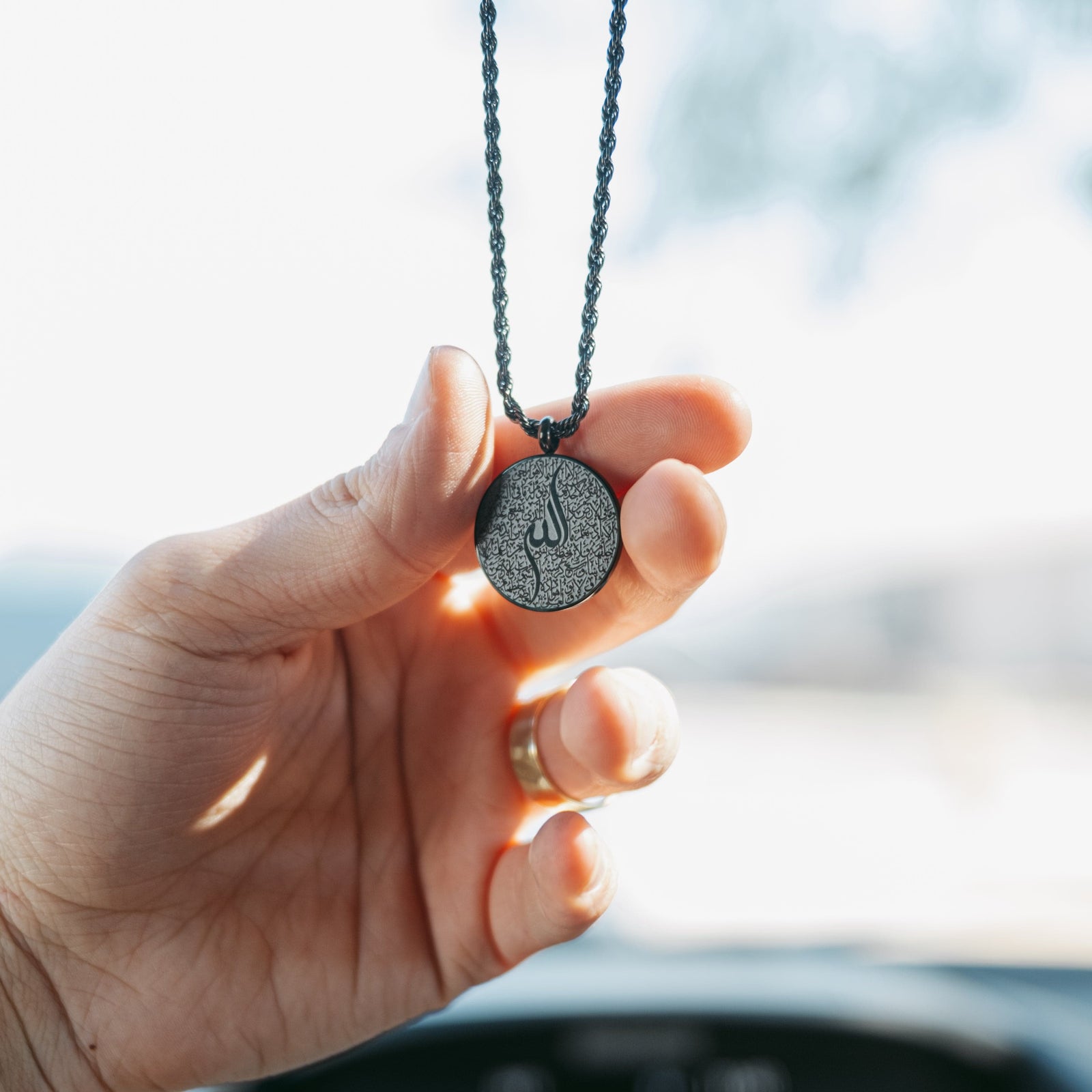 Hand holding a black Ayatul Kursi car hanger, photographed against a bright background.