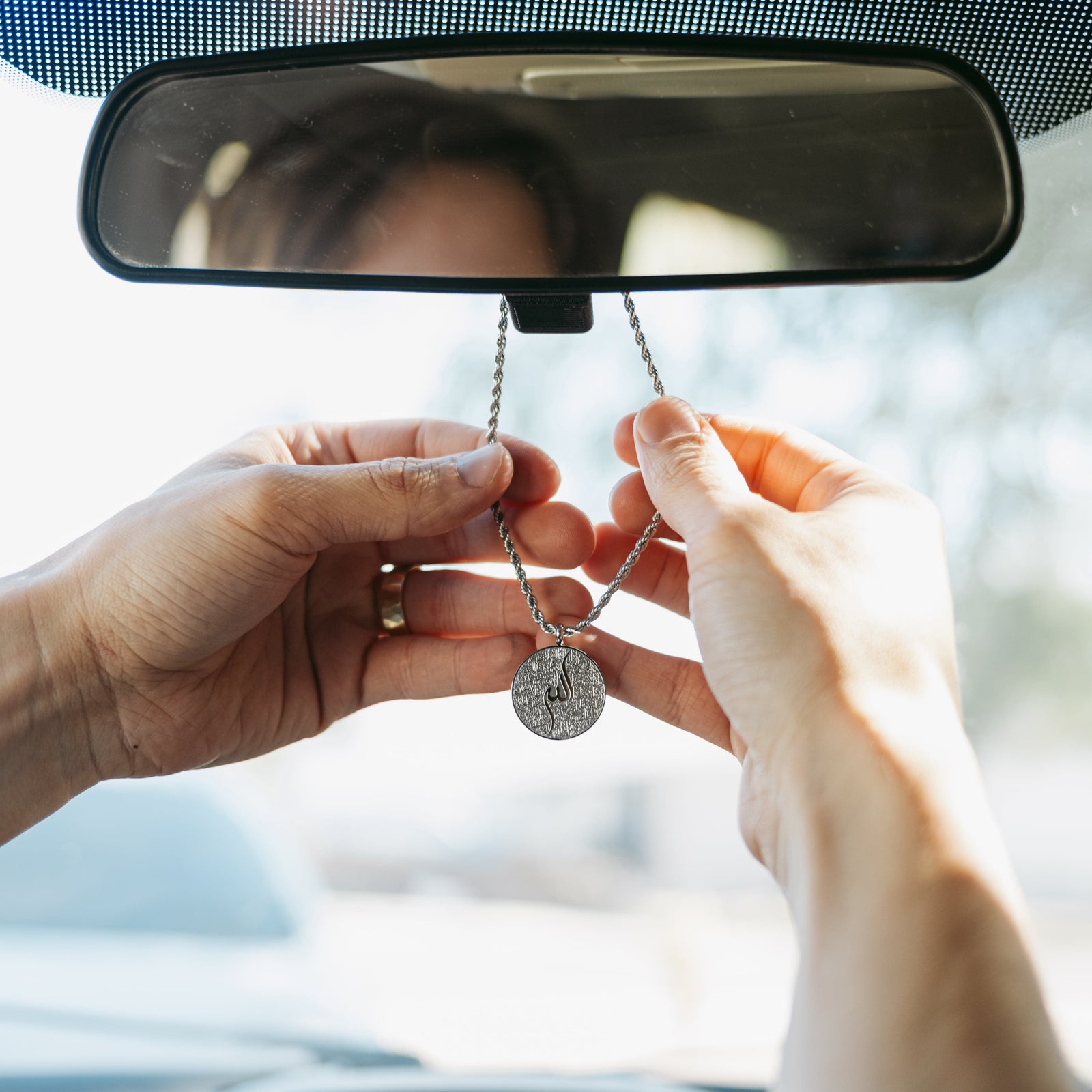 Hands adjusting a silver Ayatul Kursi car hanger on a rearview mirror inside a car.