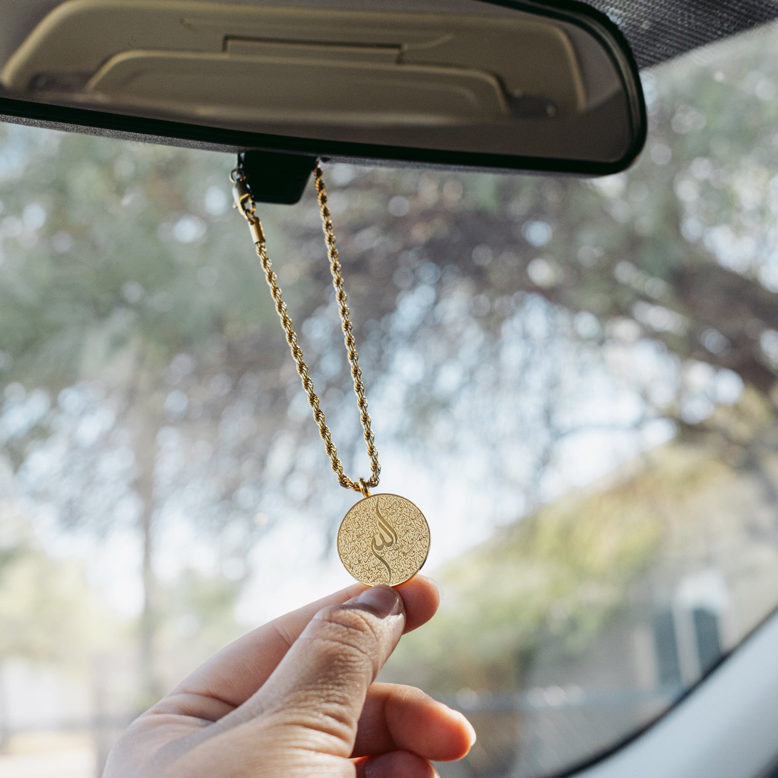 Hand holding a gold Ayatul Kursi car hanger, hanging from a rearview mirror.