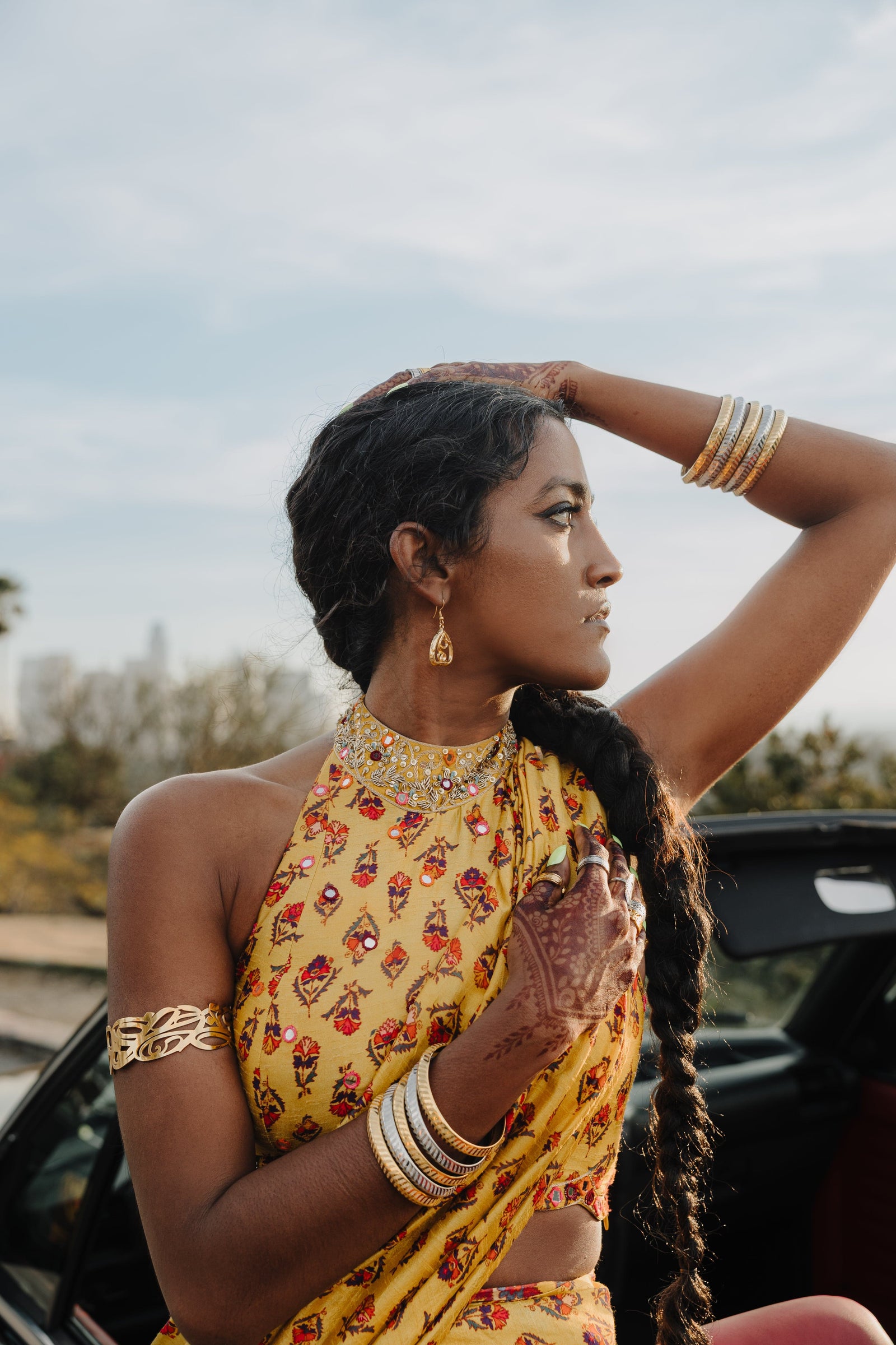 A person wearing a gold "Peepal Earring" from the Aadil x Nominal collection. The earring has a unique, abstract design with swirling lines enclosed within a roughly triangular frame. The metal is a warm gold color. The person is wearing a green top and has a hand near the earring. The image is a product shot, likely showcasing the design and craftsmanship of the earring.