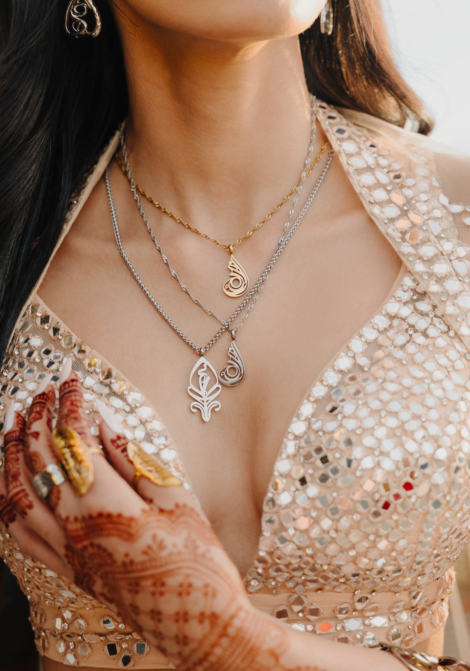 A person is wearing two delicate necklaces, one gold-plated and one silver-plated, each featuring a stylized, teardrop-shaped pendant with intricate cut-out details. The necklaces are worn against a light beige top. A hand with henna designs and gold rings is visible.