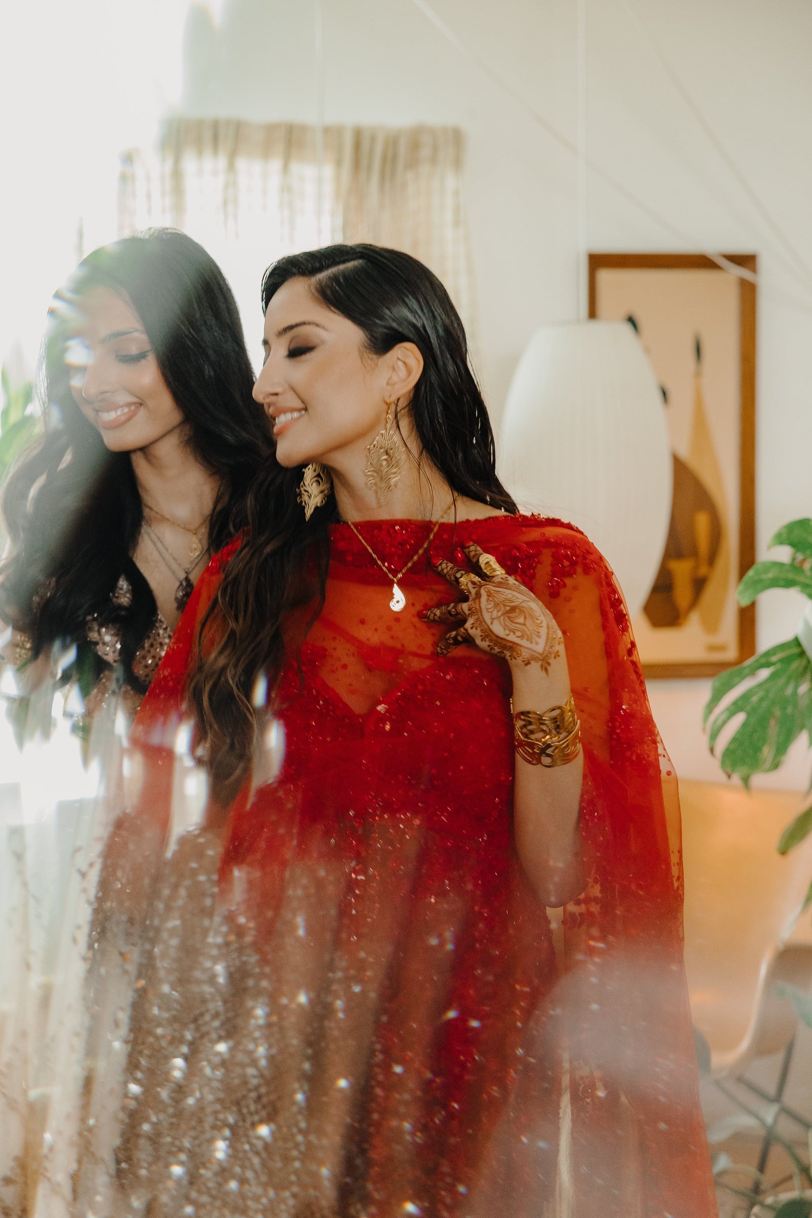 A delicate gold-plated chain necklace featuring a dainty, approximately 2 cm teardrop pendant with intricate, swirling cut-out details and a shiny finish. The necklace is worn by a person with dark hair and is shown against a blurred red, semi-sheer garment. A hand with henna designs and gold bangles is gently touching the pendant. Another person with dark hair is partially visible in the background, slightly out of focus.