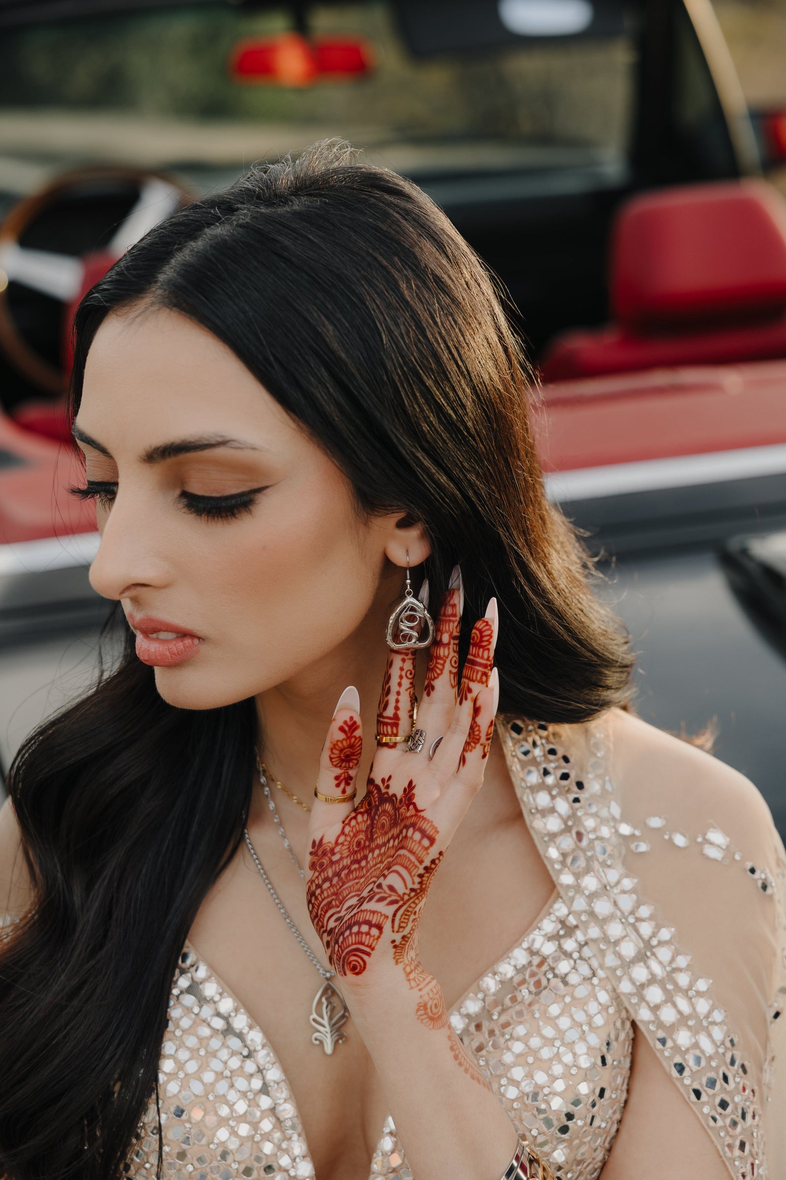 A person with long, dark hair is shown in profile, looking down and to the left. They are wearing silver "Peepal Earrings" from the Aadil x Nominal collection. The earrings have a unique, abstract design with swirling lines enclosed within a roughly triangular frame. The person is also wearing a silver necklace and a light-colored garment with mirror embellishments. Their hand, adorned with intricate henna patterns, is near their face. The background shows part of a red car interior and a blurred outdoor sc