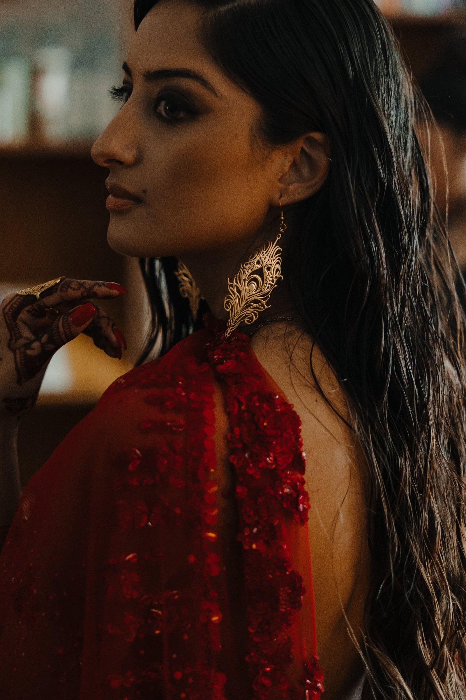 A person with long, dark, slightly wavy hair is shown in profile, facing left. They are wearing a gold "Peacock Feather Earring" from the Aadil x Nominal collection. The earring is intricately designed in the shape of a peacock feather with detailed cut-out patterns. The person is also wearing a red garment with embellishments and has henna on their hands. The background is softly blurred, suggesting an indoor setting.