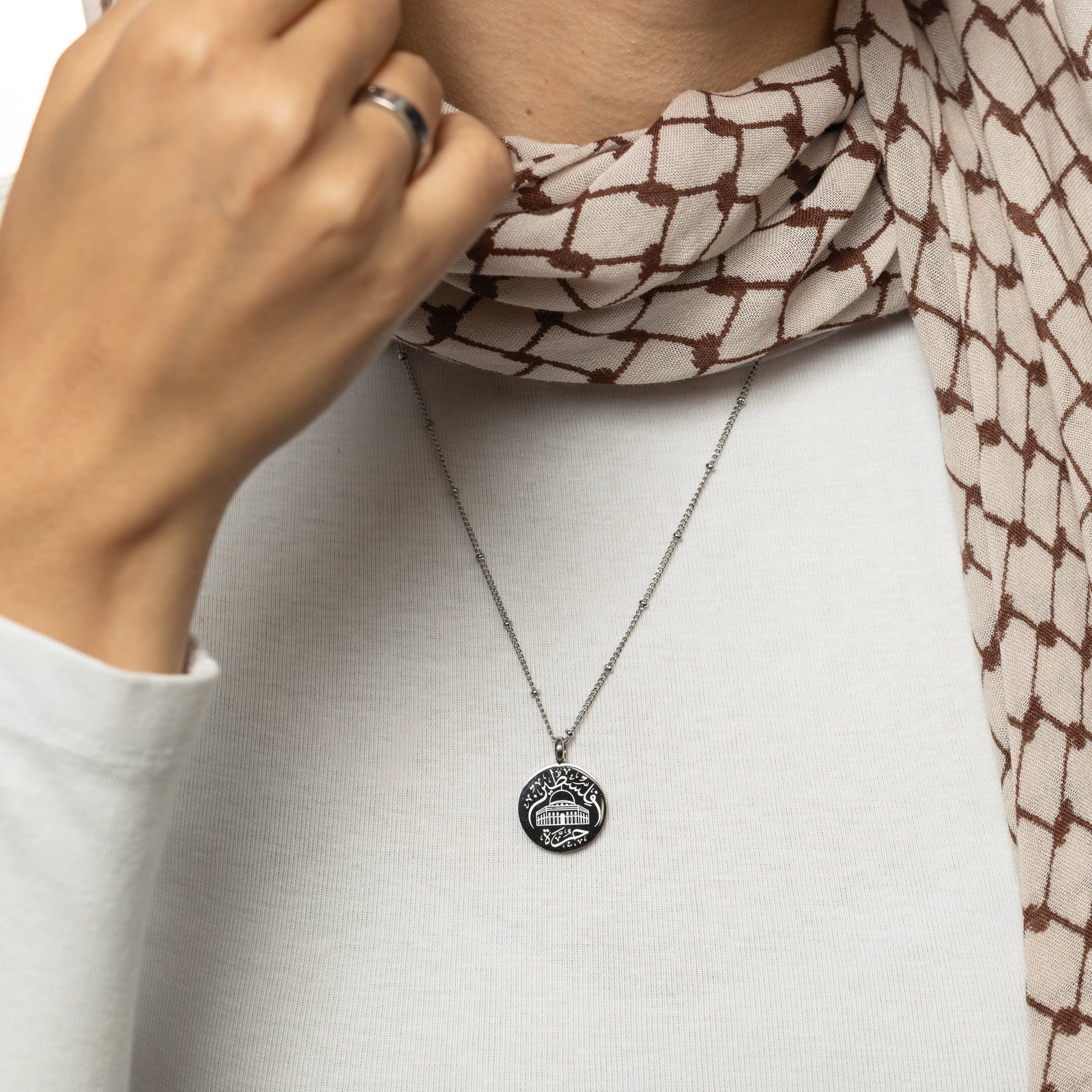 A woman wearing a white shirt, keffiyeh patterned scarf, and a silver Al-Aqsa ‘Free Palestine’ Necklace