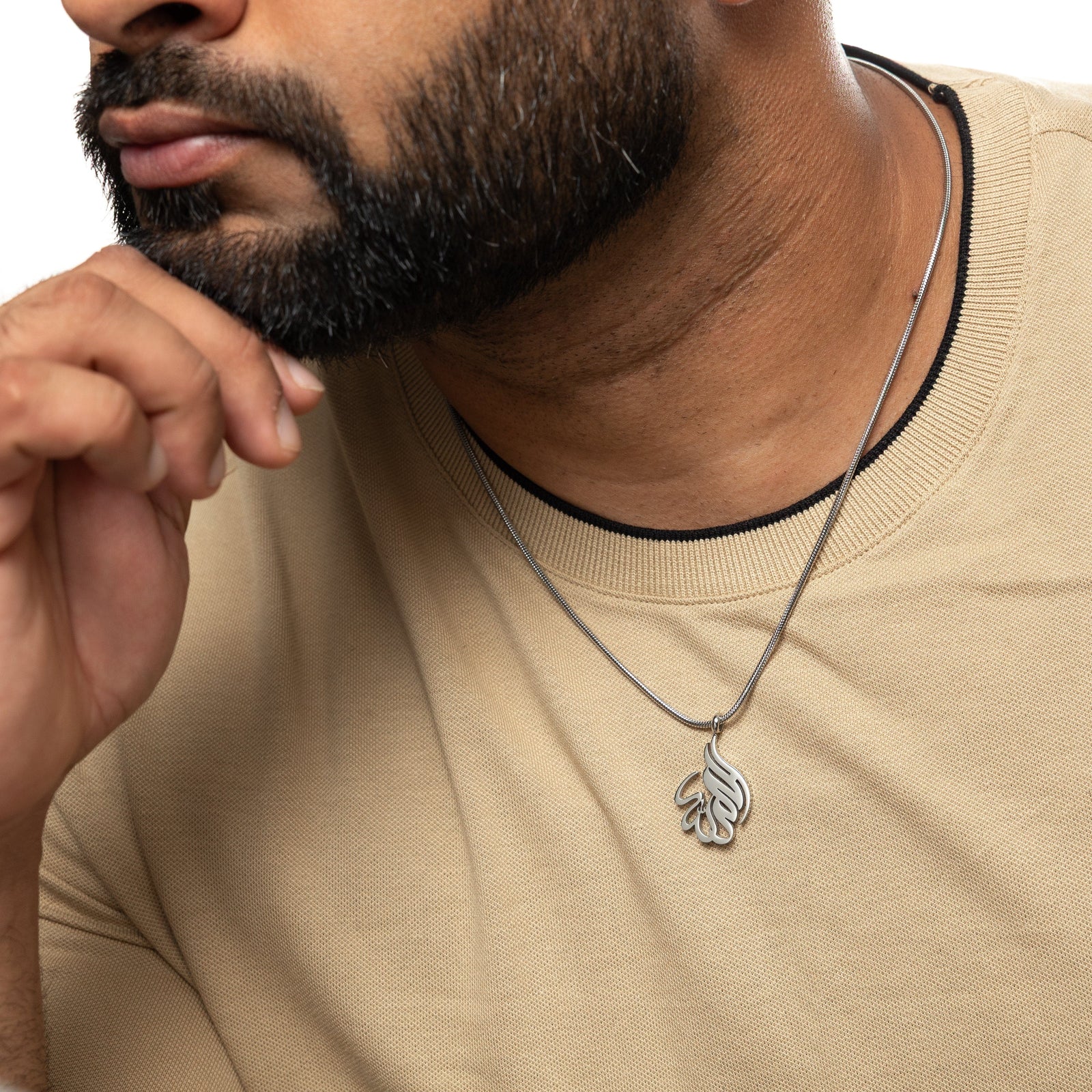 Man wearing a silver 'Alhamdulillah' calligraphy necklace with a beige shirt, resting hand on chin.