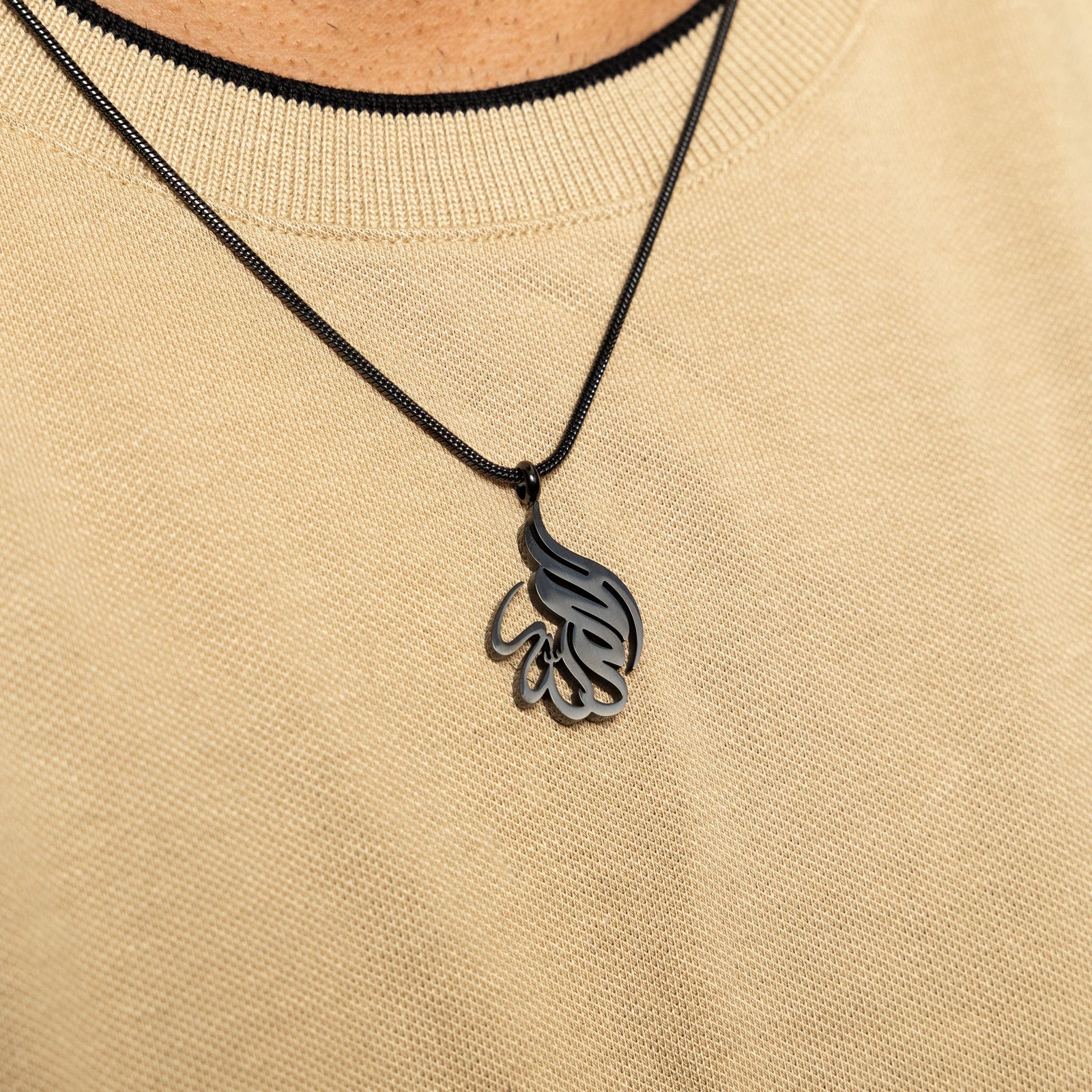Black 'Alhamdulillah' calligraphy necklace worn over a beige textured top.