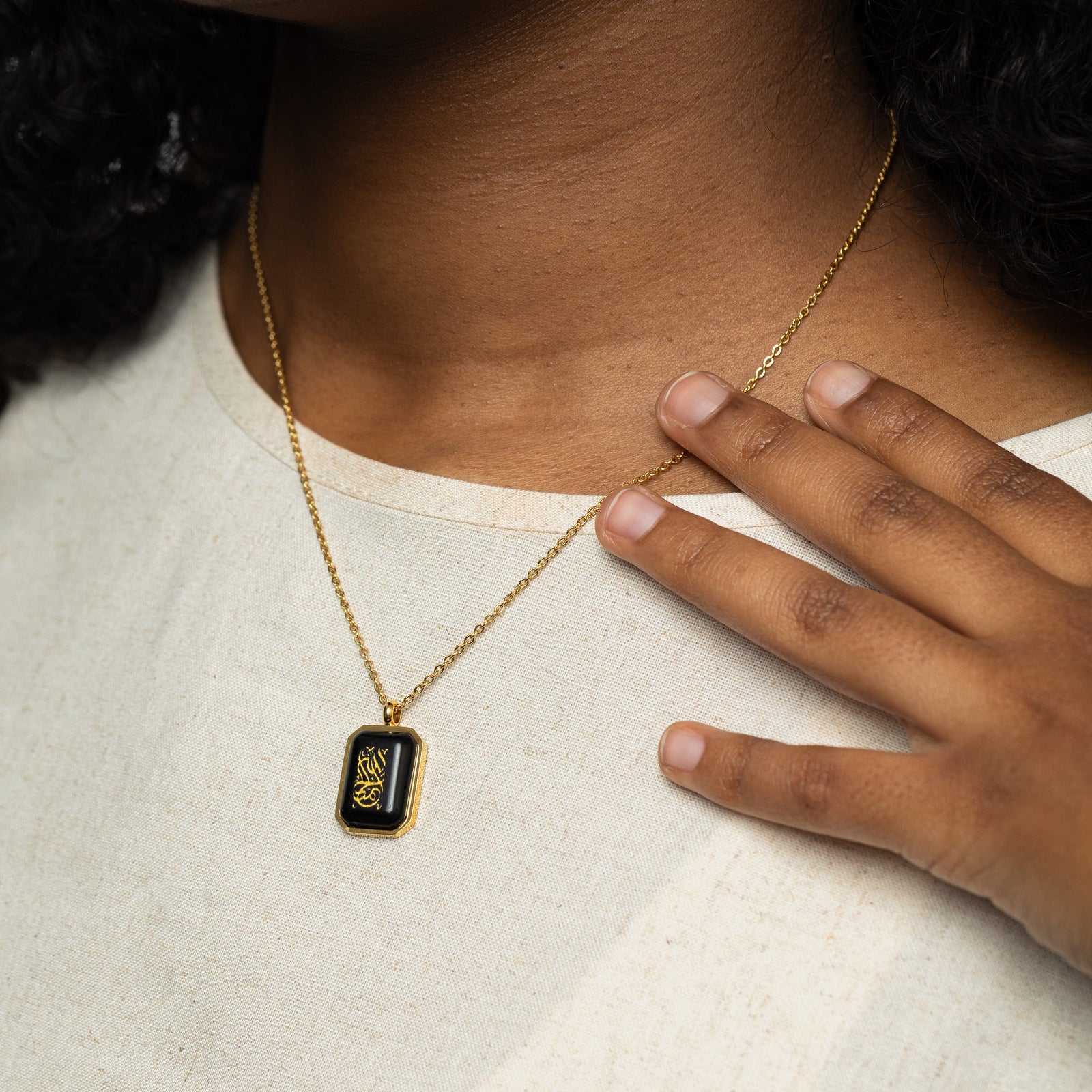 Close-up of a woman wearing a gold chain necklace with a rectangular black obsidian pendant engraved with "Alhamdulillah" in Arabic calligraphy, hand lightly touching the necklace chain over a cream shirt.