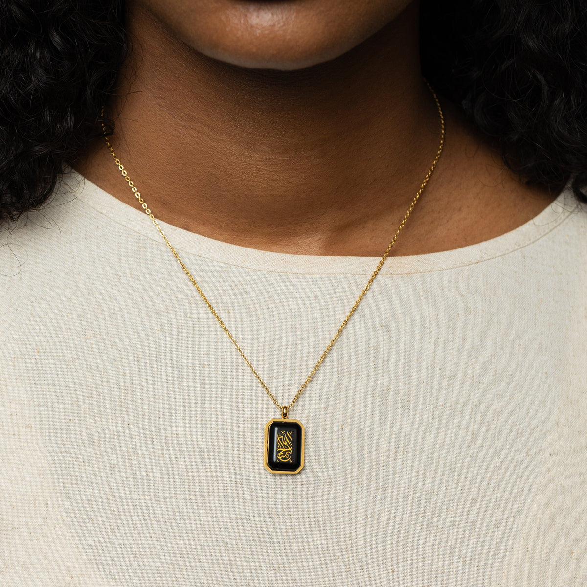 Woman wearing a gold chain necklace with a rectangular black obsidian pendant engraved with "Alhamdulillah" in Arabic calligraphy, styled with a cream shirt.