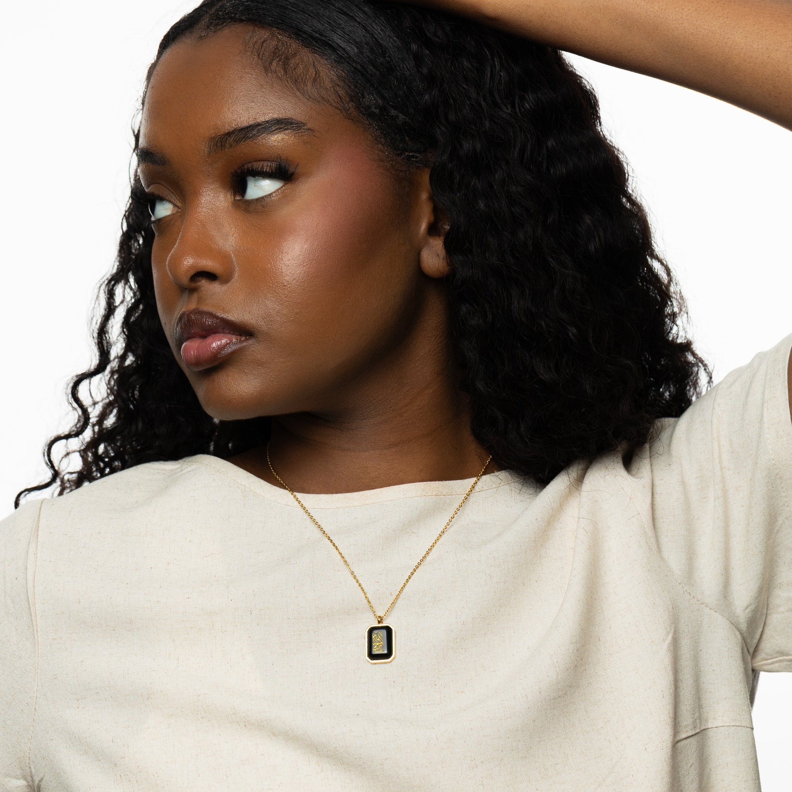 Woman wearing a gold chain necklace with a rectangular black obsidian pendant engraved with "Alhamdulillah" in Arabic calligraphy, styled with a cream top.