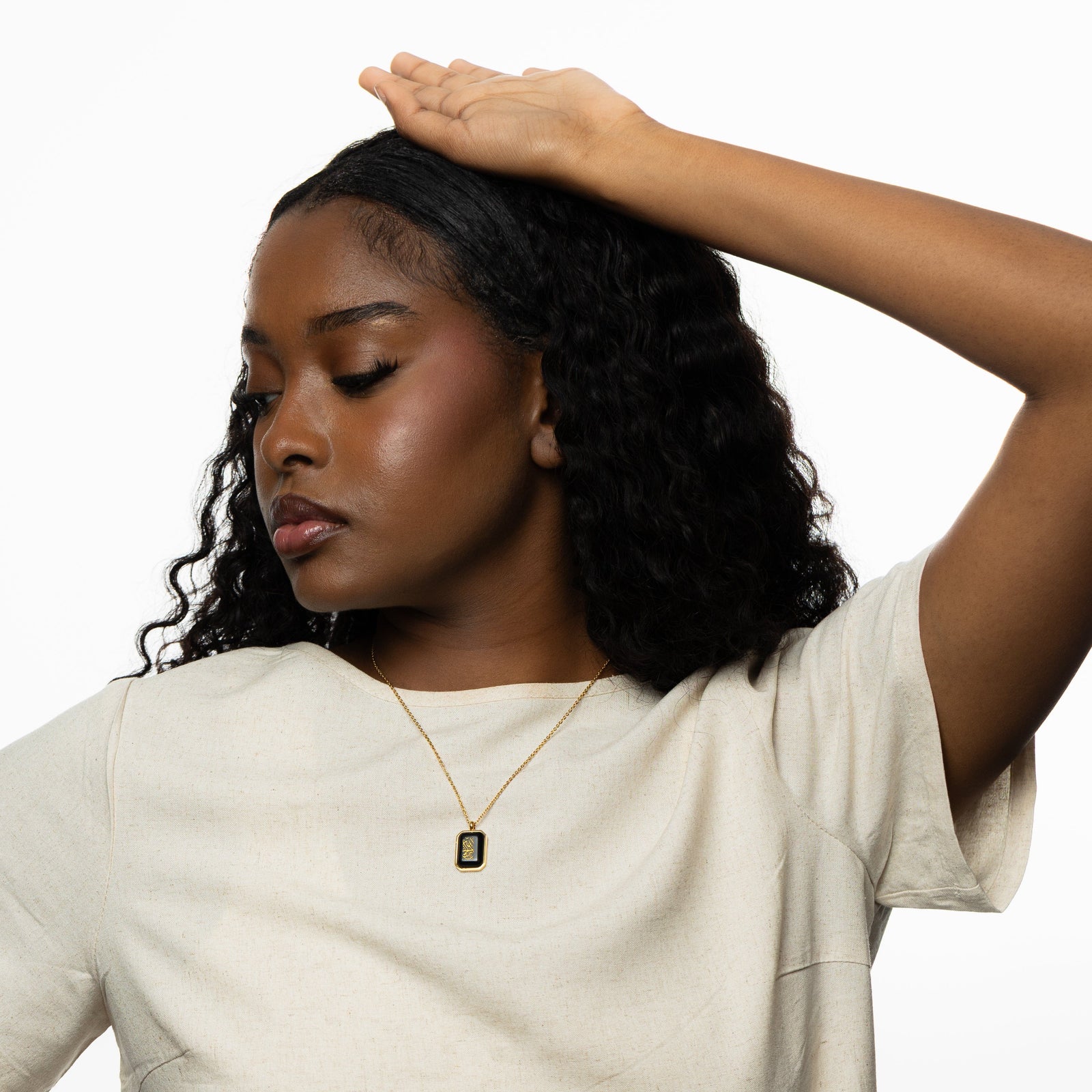 Woman wearing a gold chain necklace with a rectangular black obsidian pendant engraved in Arabic calligraphy reading "Alhamdulillah," styled with a cream shirt, arm raised above head.