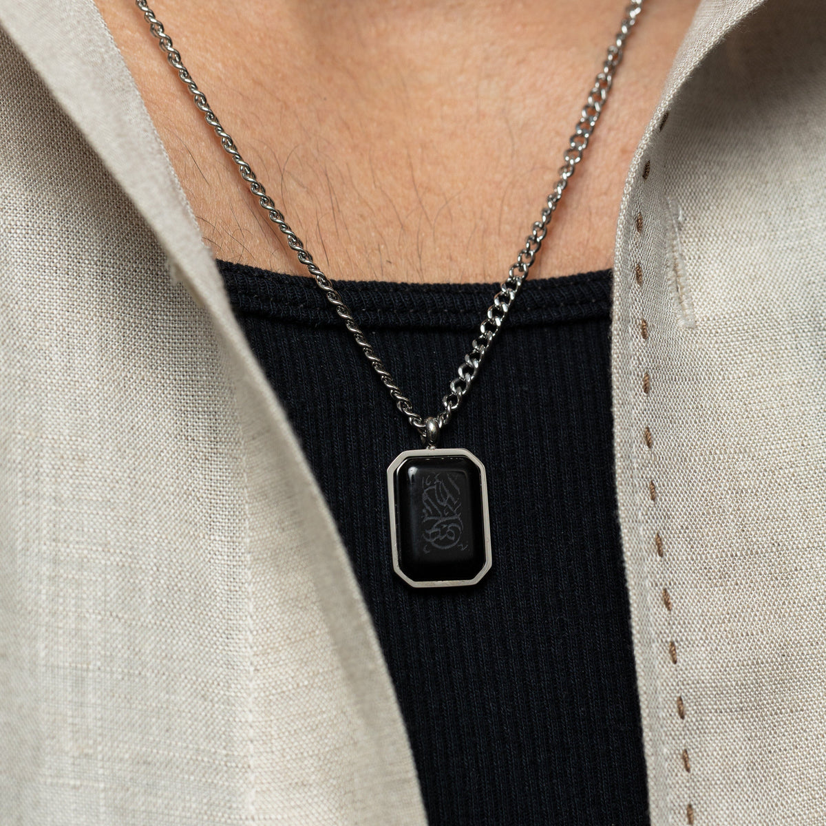 Man wearing a silver chain necklace with a rectangular black obsidian pendant engraved with "Alhamdulillah" in Arabic calligraphy, styled with a black ribbed shirt and cream open shirt layered on top.