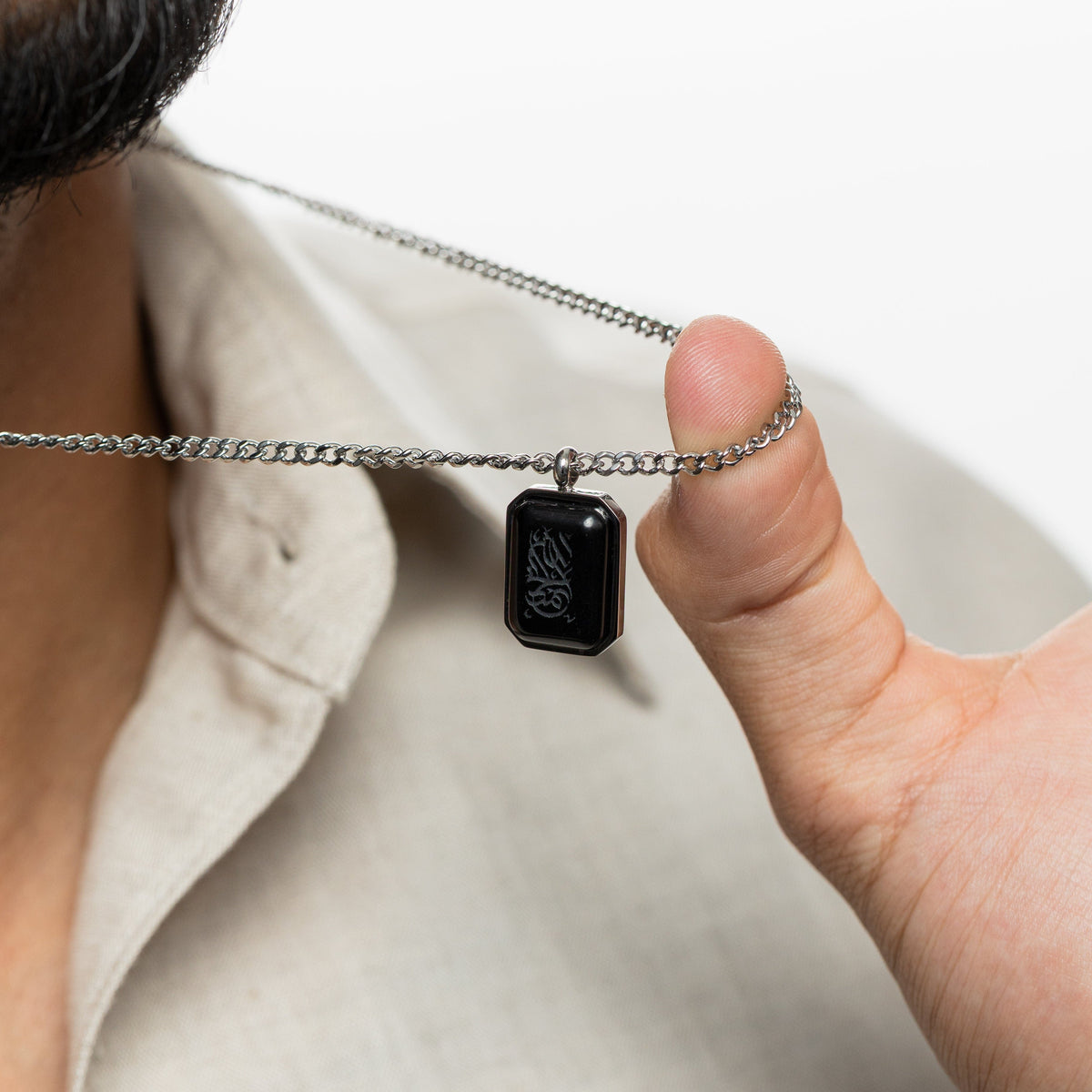 Man holding up a silver chain necklace with a rectangular black obsidian pendant engraved with "Alhamdulillah" in Arabic calligraphy.