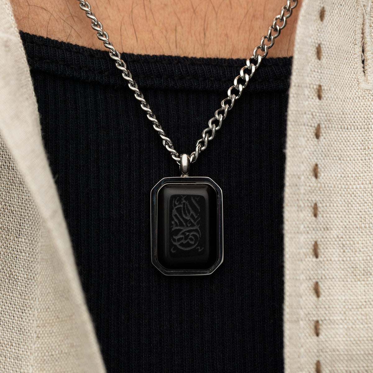 Close-up of a man wearing a silver chain necklace with a rectangular black obsidian pendant engraved in Arabic calligraphy reading "Alhamdulillah".