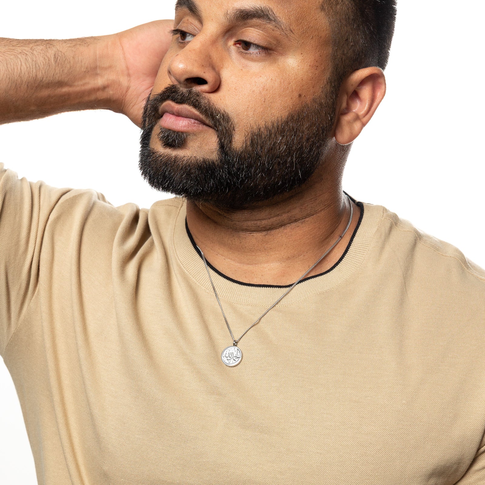 Man wearing a silver Alhamdulillah necklace, styled with a beige textured shirt, hand resting behind his head.