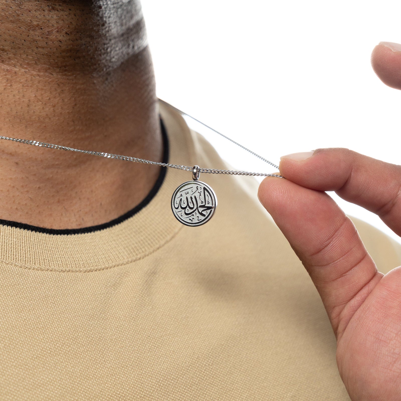 Close-up of a man holding up a silver necklace with a circular pendant engraved with 'Alhamdulillah' calligraphy, worn over a beige shirt.