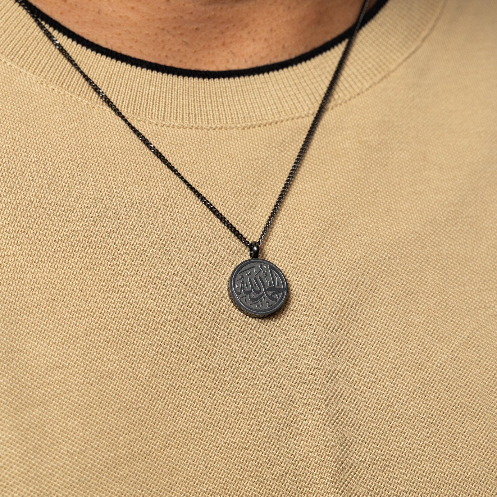 Black Alhamdulillah token necklace worn over a beige textured shirt.