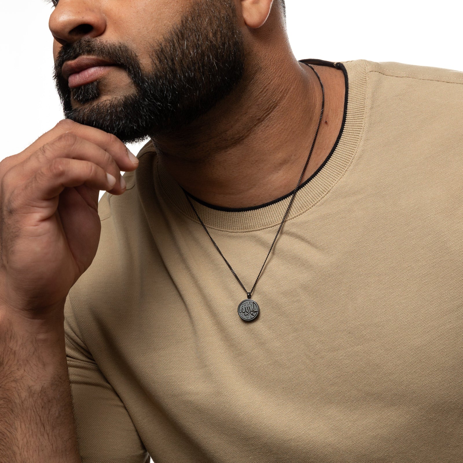 Man wearing a black necklace with a circular 'Alhamdulillah' calligraphy pendant, styled with a beige shirt, hand resting on chin