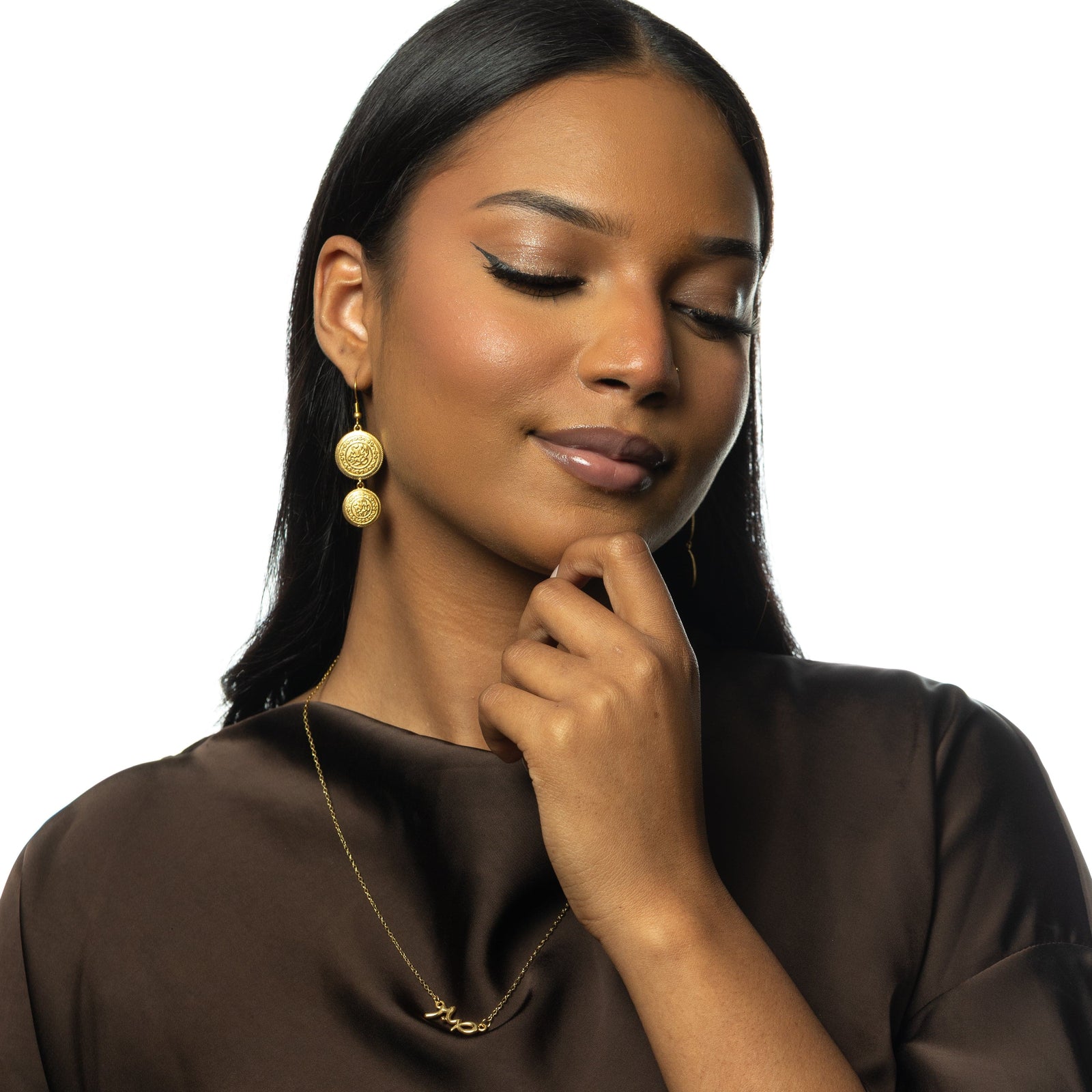 Photo of the model with one of her hands is gently raised to her chin, with her fingers lightly touching it, giving the impression that she is in a thoughtful or reflective moment. Her accessories include gold Alhamdulillah Coin Earrings and a delicate gold necklace with a small script-style pendant.