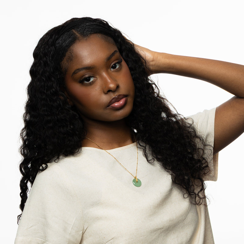 Woman wearing the green aventurine stone pendant necklace with a gold chain pimage_ styled against a neutral top pimage_ one arm lifted toward her hair.