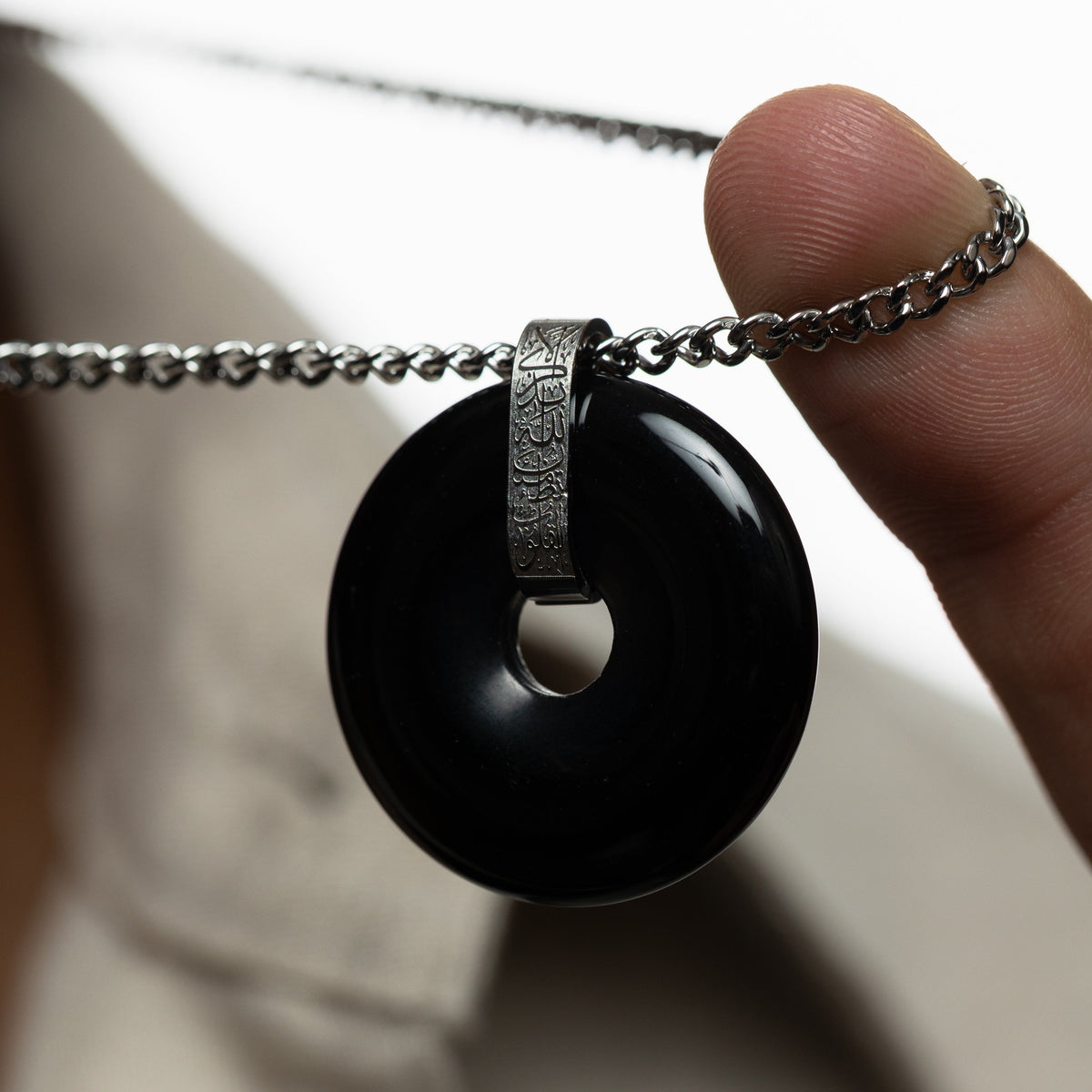 Hand holding the chain of the necklace, highlighting the glossy black onyx pendant with silver bail engraved in Arabic calligraphy.