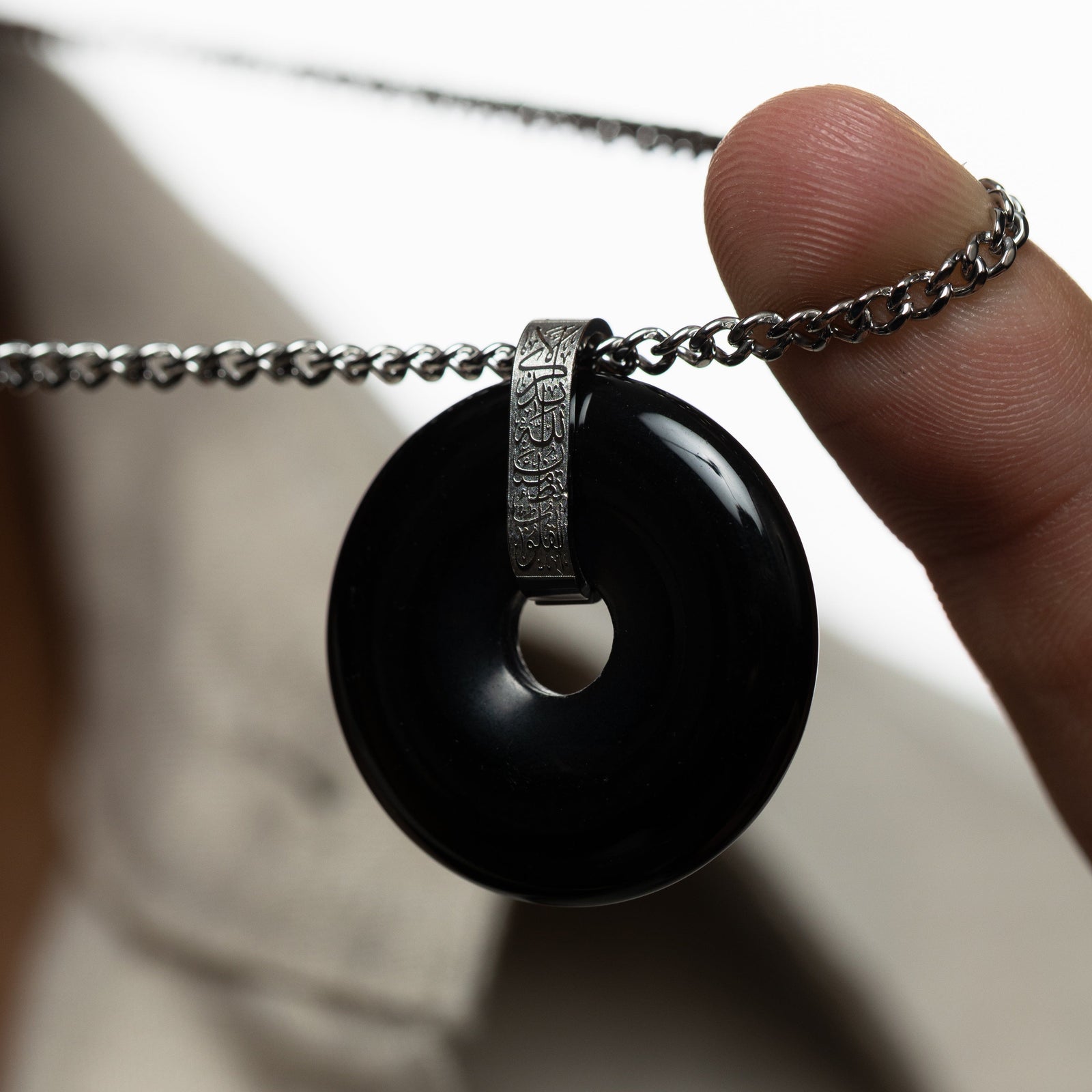Hand holding the chain of the necklace, highlighting the glossy black onyx pendant with silver bail engraved in Arabic calligraphy.