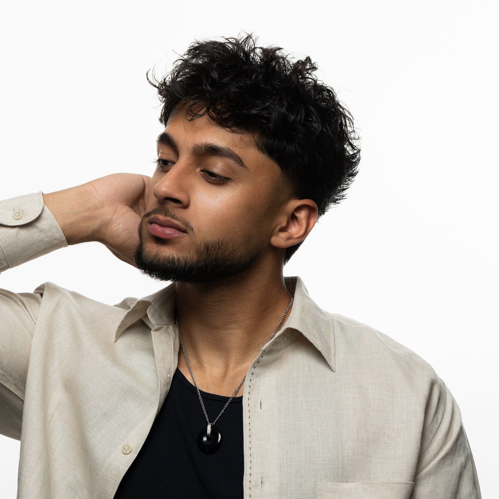 Man wearing the black onyx stone necklace on a silver chain, styled with a black shirt and beige open button-down, hand resting behind his head.