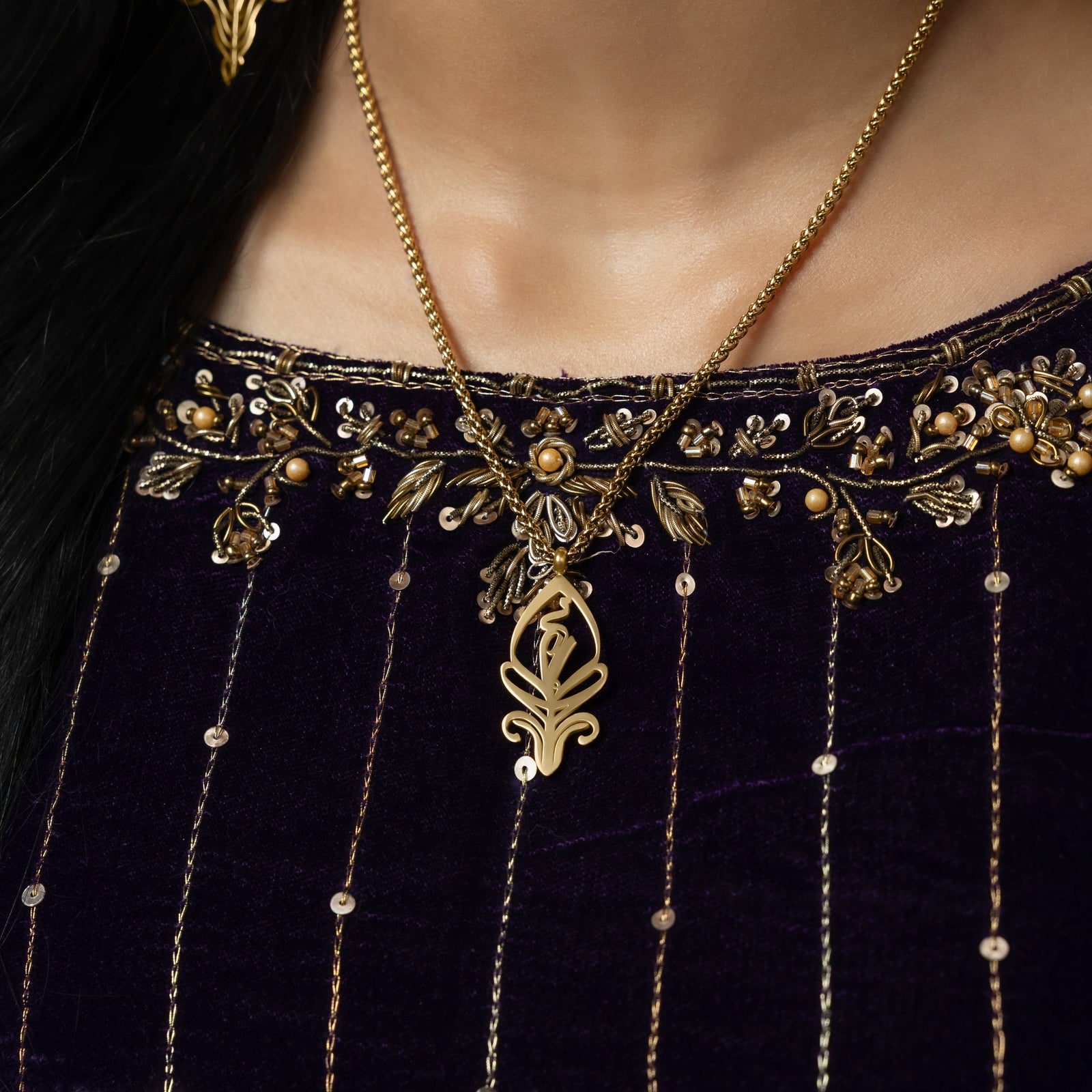 A delicate gold-plated chain necklace featuring a stylized, teardrop-shaped pendant with intricate cut-out details. The necklace is worn by a person with dark hair and is shown against a dark blue velvet garment with gold embroidery.