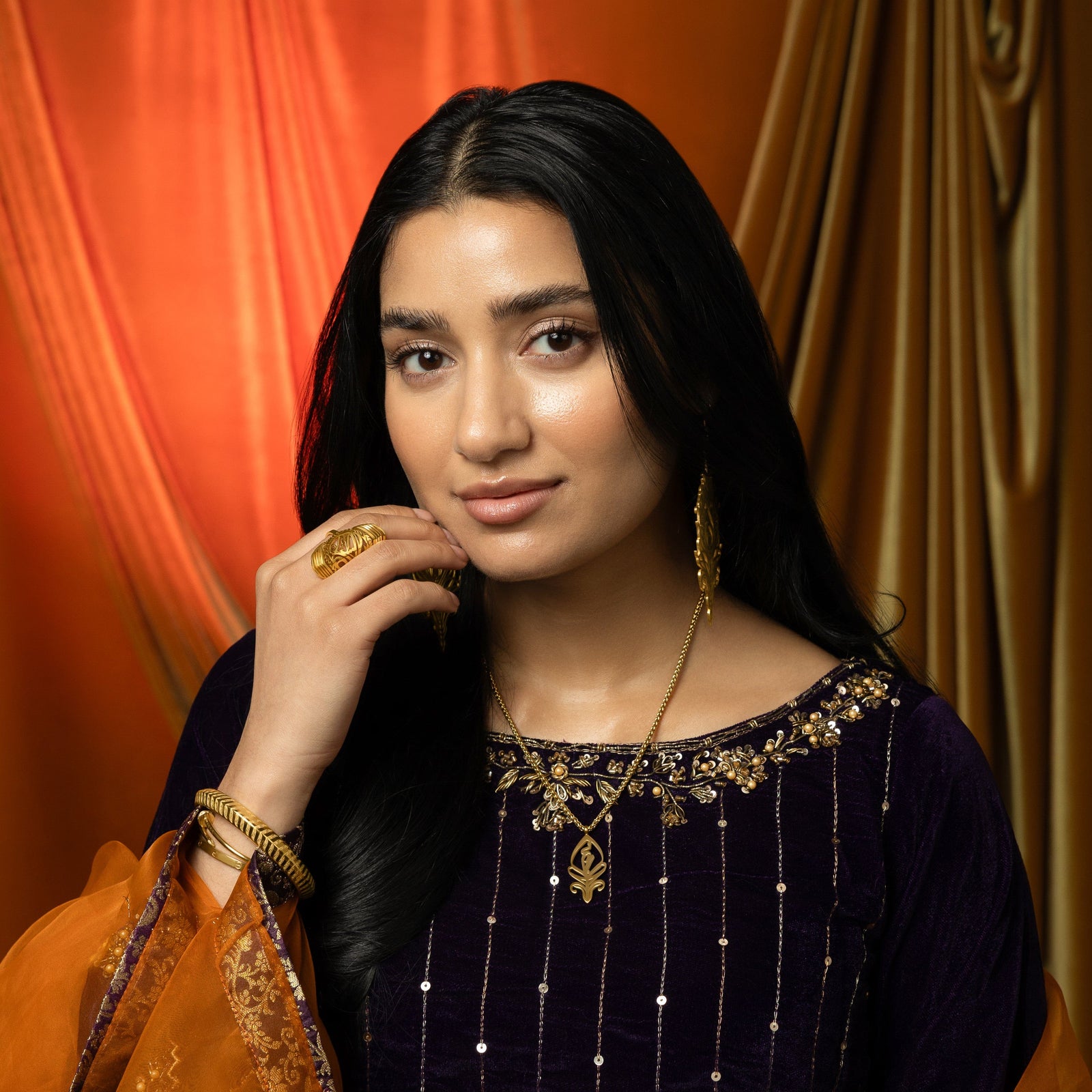A delicate gold-plated chain necklace featuring a stylized, teardrop-shaped pendant with intricate cut-out details. The necklace is worn by a person with dark hair and is shown against a dark blue velvet garment with gold embroidery.