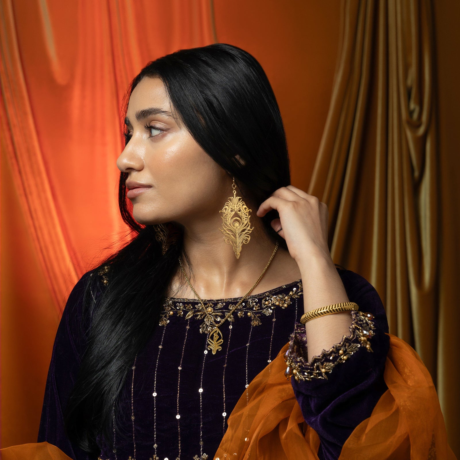 A delicate gold-plated chain necklace featuring a stylized, teardrop-shaped pendant with intricate cut-out details. The necklace is worn by a person with dark hair and is shown against a blurred background. A hand with henna designs and gold rings is gently touching the pendant.