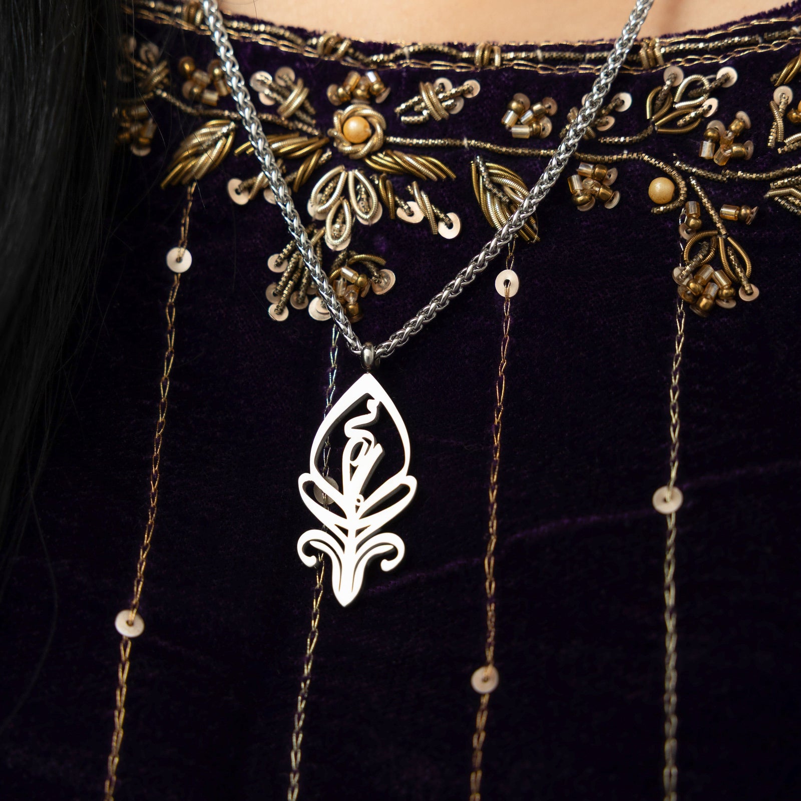 A delicate silver-plated chain necklace featuring a stylized, teardrop-shaped pendant with intricate cut-out details. The necklace is worn by a person with dark hair and is shown against a dark blue velvet garment with gold embroidery.