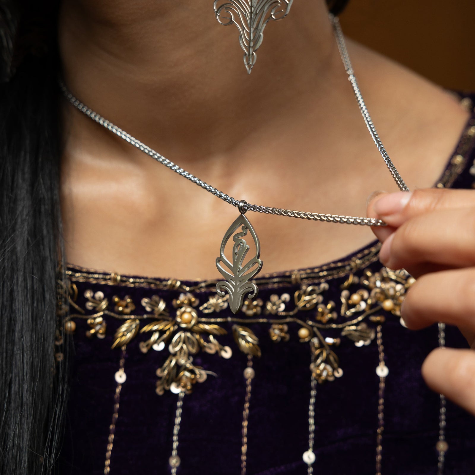 A delicate silver-plated chain necklace featuring a stylized, oval pendant with intricate cut-out details. The necklace is worn by a person with dark hair and is shown against a dark blue velvet garment with gold embroidery.