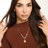 Woman wearing an 18k gold plated necklace with a detailed daisy flower pendant and translucent white tube beads, paired with matching drop earrings