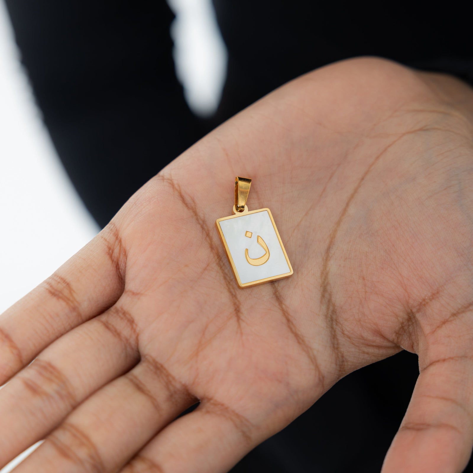 Close-up of the Arabic letter charm "ن" (Noon) in 18K gold plating, with an iridescent pearl background.