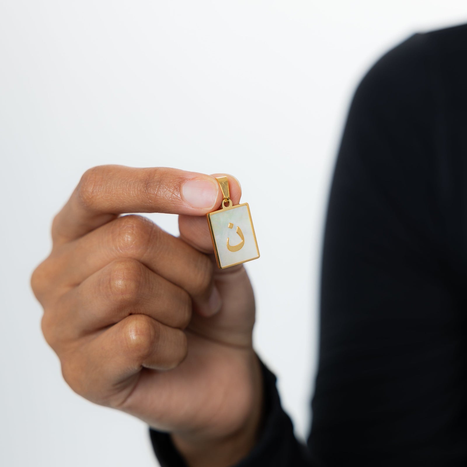 Close-up of the Arabic letter charm "ن" (Noon) in 18K gold plating, with an iridescent pearl background.
