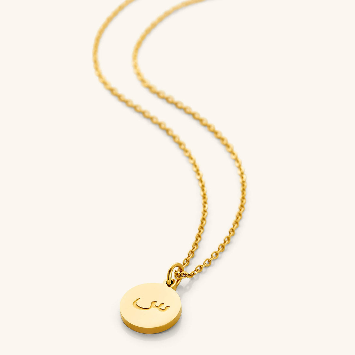 An 18k gold plated necklace with a round pendant engraved with Arabic Letter on a light background.