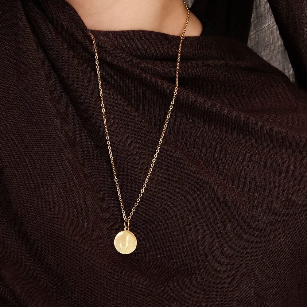 Close up of the gold Arabic Letter Necklace against the brown fabric of the model's clothing.