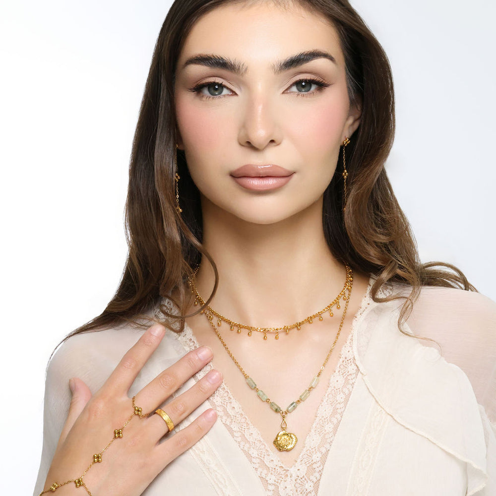 Woman wearing an 18k gold plated layered necklace with a poppy flower pendant and pale green tube beads, paired with drop earrings and a matching hand chain.