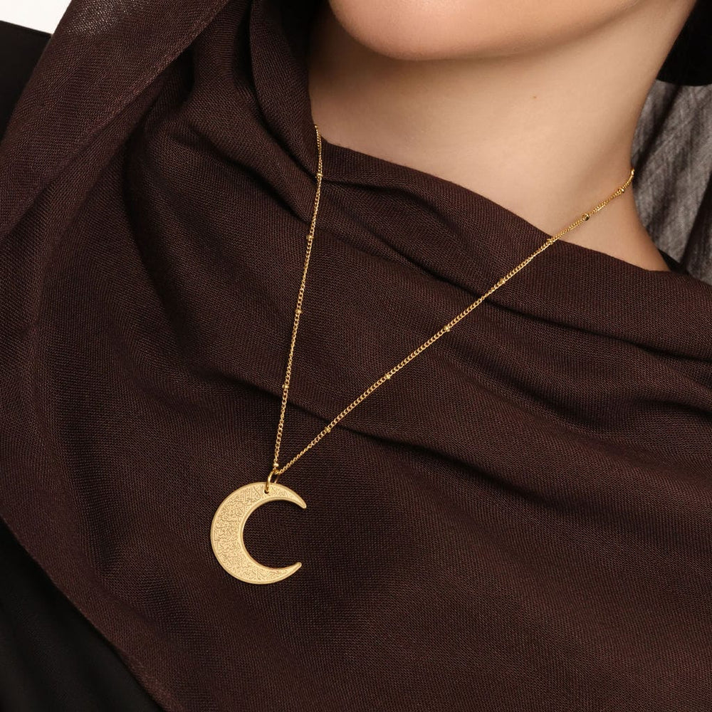 Close up of the gold Ayatul Kursi Crescent Necklace against the brown fabric of the model's clothing.