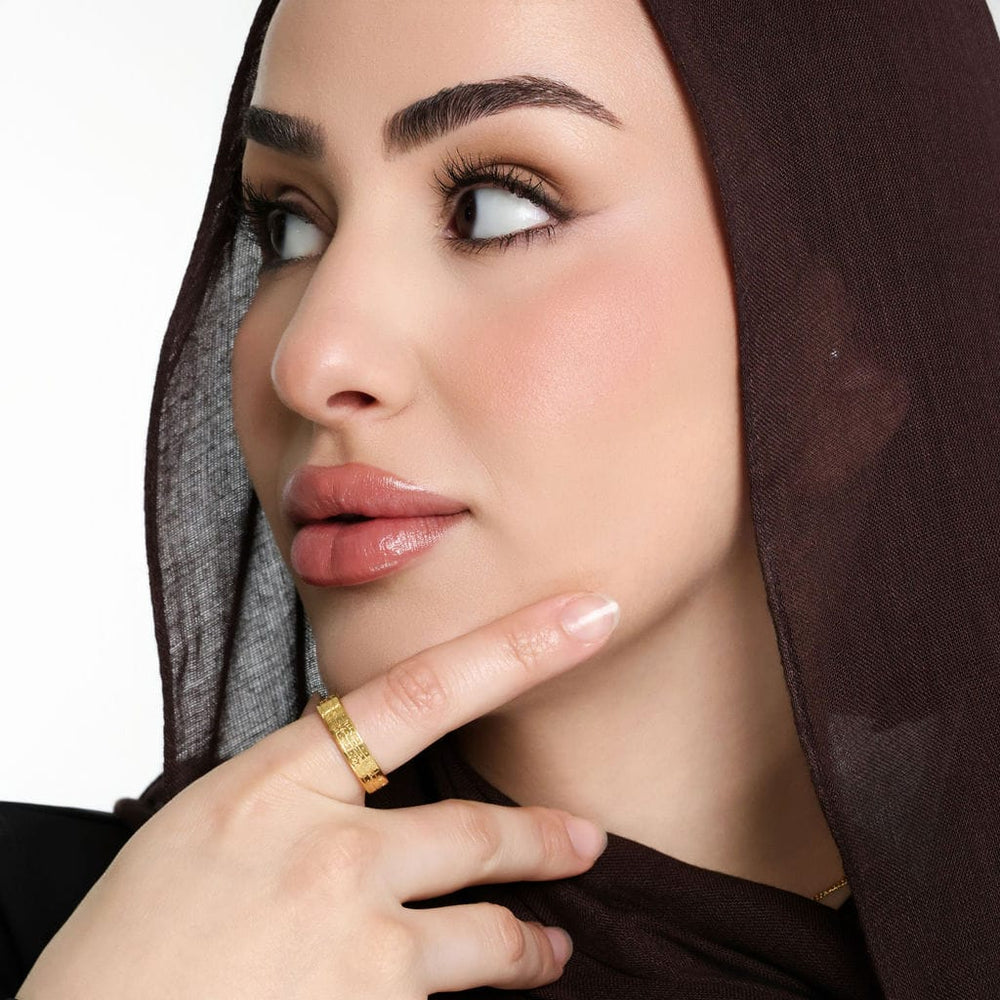 Close up of the model's face while the model looks to their side, away from the camera. The model touches their chin with their right index finger to show the gold Ayatul Kursi Ring | Women they are wearing.