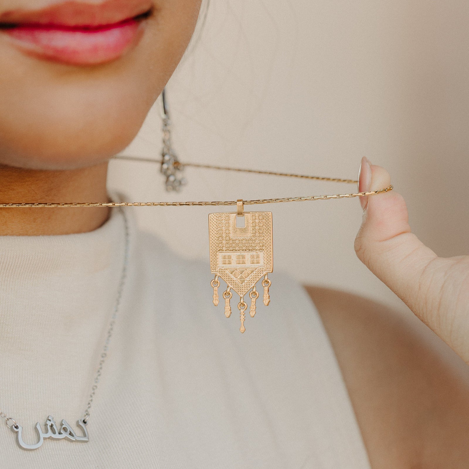 Close-up of the Raining Tatreez Necklace being held up by the chain, focusing on the textured Tatreez pendant and the five dangling charms.