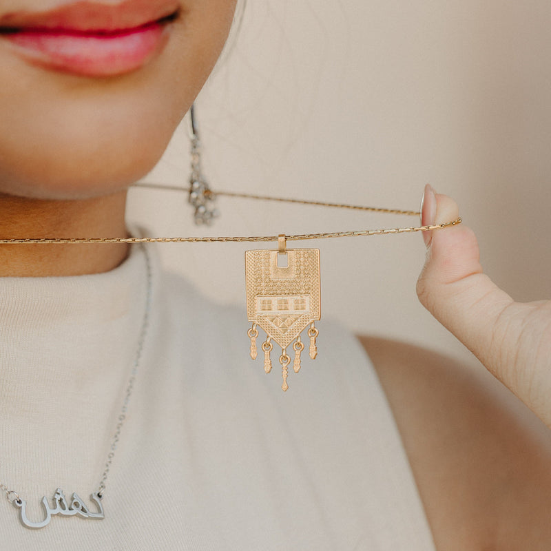 Close-up of the Raining Tatreez Necklace being held up by the chain pimage_ focusing on the textured Tatreez pendant and the five dangling charms.