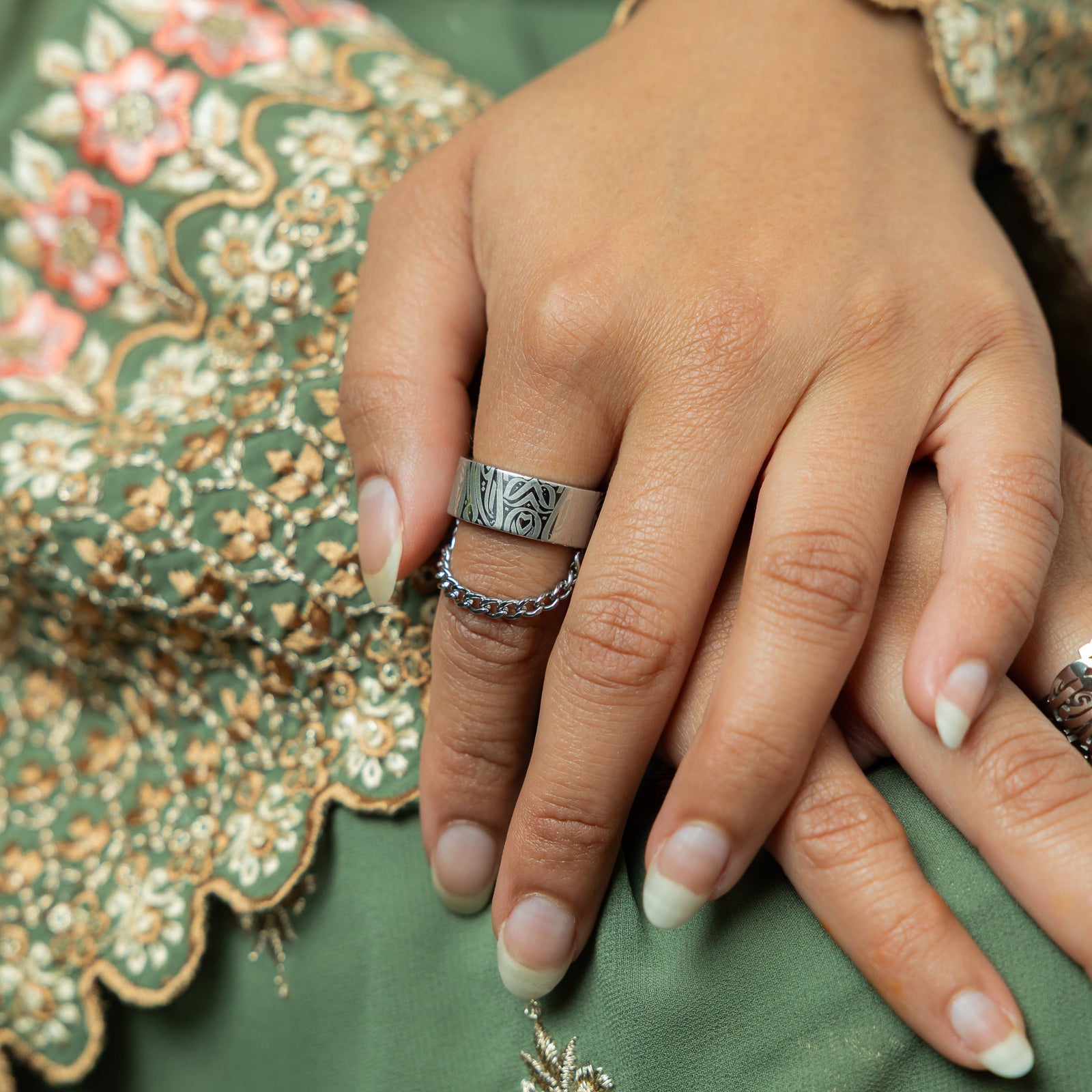 A wide, silver-plated ring with a rectangular face and intricate, abstract designs engraved across the surface. A delicate silver chain is attached to either side of the ring. The ring is worn on a woman's finger.