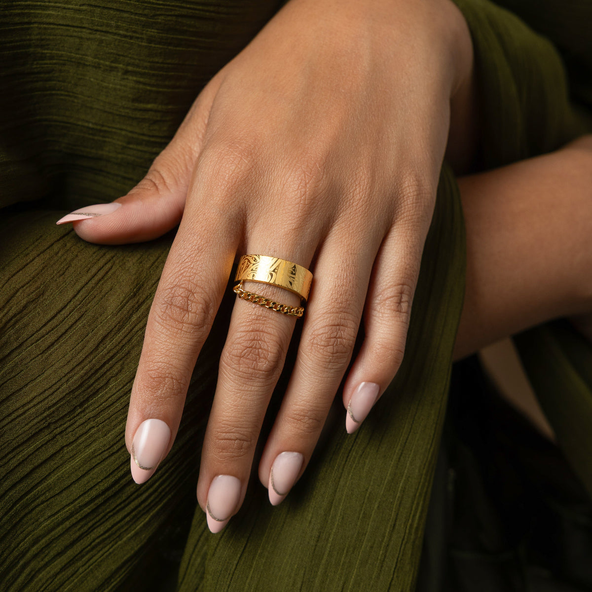 A wide, gold-plated ring with a rectangular face and intricate, arabic calligraphy designs engraved across the surface. The ring is worn on a person's finger.
