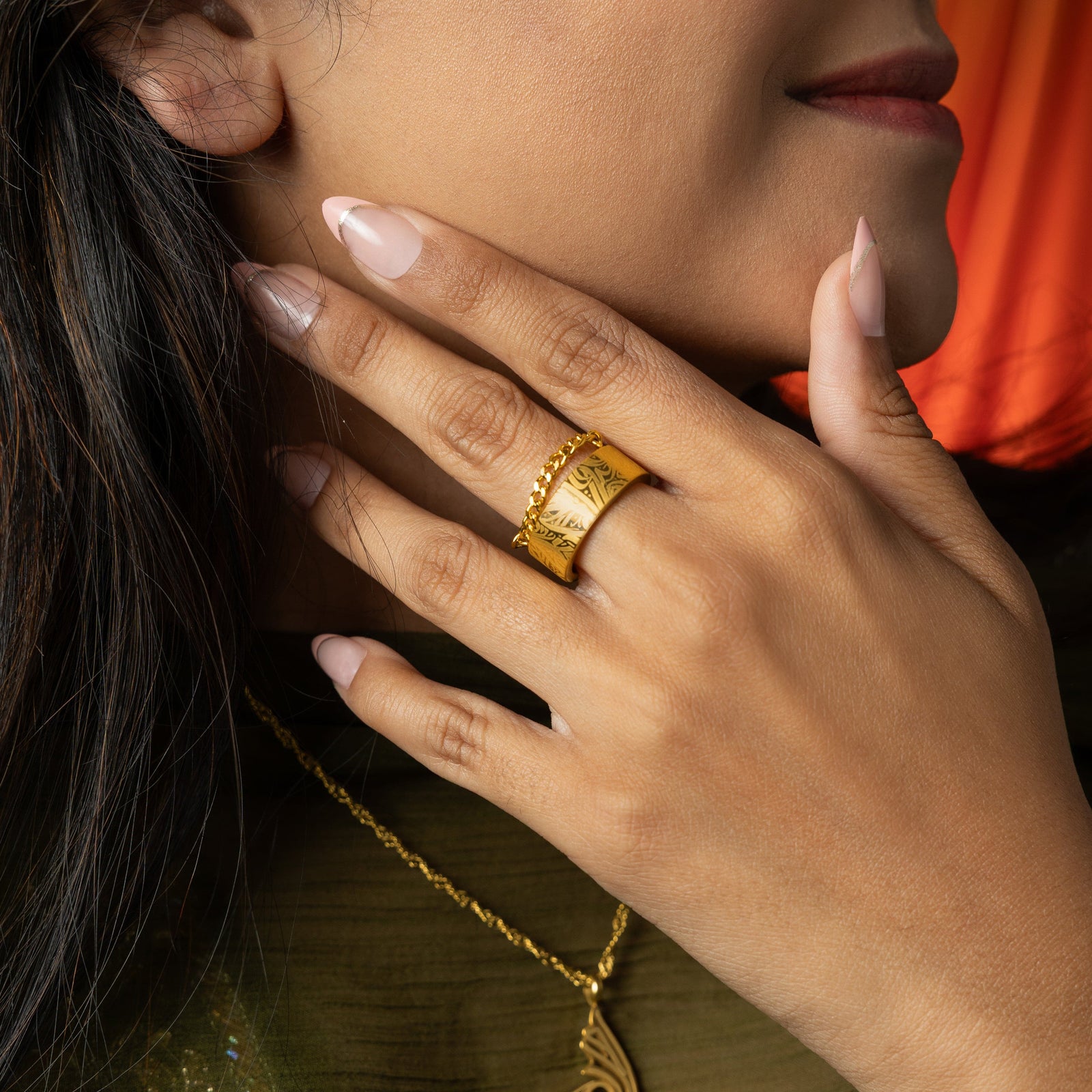 A wide, gold-plated ring with a rectangular face and intricate, calligraphy engraved across the surface worn by a woman on her middle finger.
